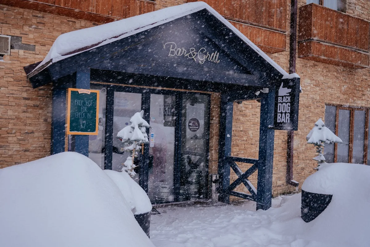 The snowed-in wooden porch and hanging sign of Black Dog Bar in Gudauri during a snowfall