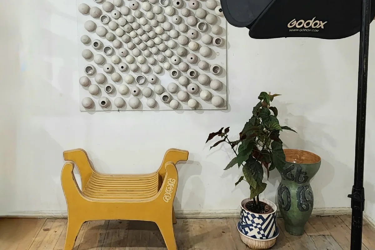 A wall piece of pale clay domes above a yellow wooden bench, with a potted plant and a green glazed vase, inside the Ceramicart studio