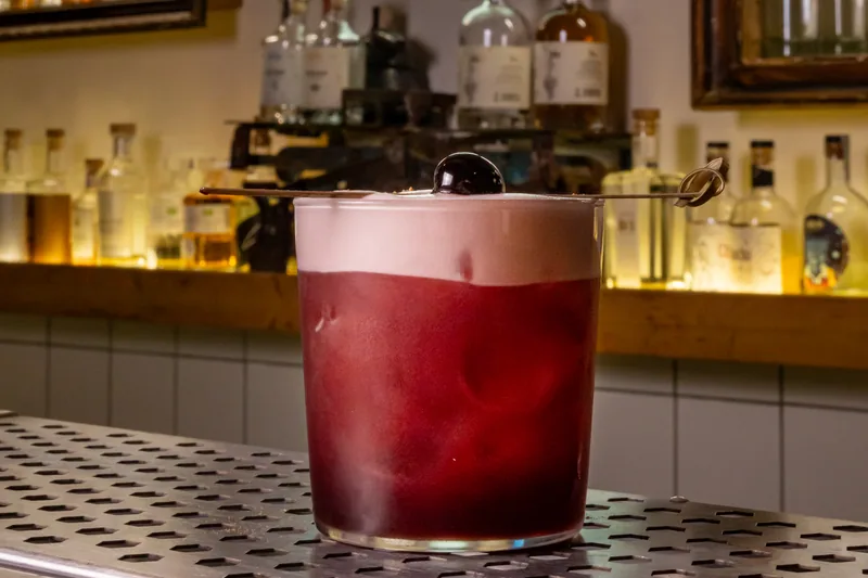 A red sour cocktail with foam and a cherry on the bar at Chacha Time, with chacha bottles behind