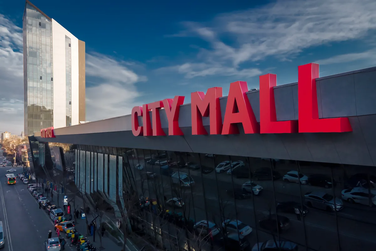The big red City Mall sign over the glass front of City Mall Saburtalo, with a tower block behind