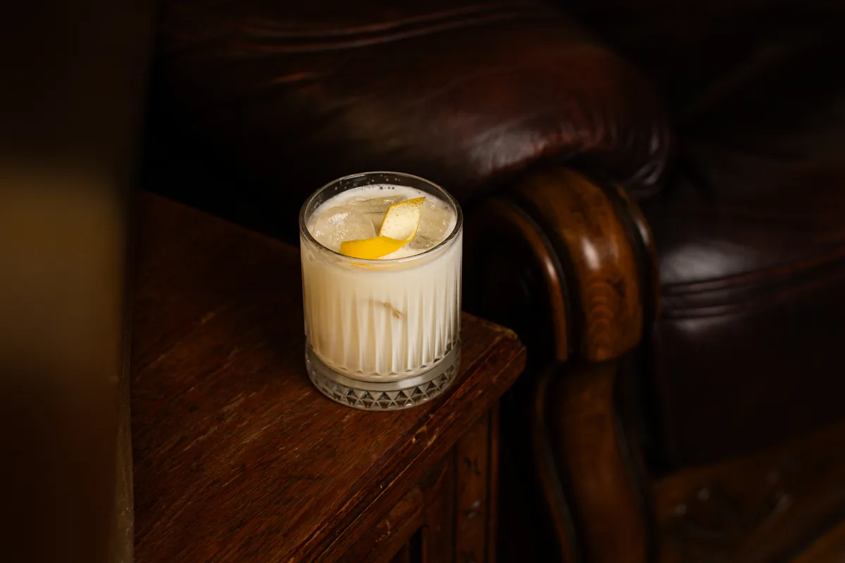 Pale creamy cocktail on ice with a lemon twist in a cut-crystal rocks glass, beside a leather armchair at Crossroads Bar