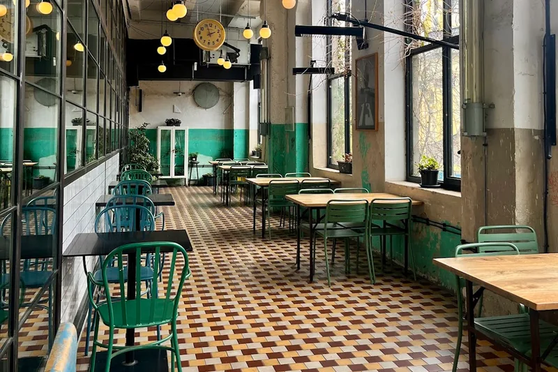 Long dining hall with a checkered tile floor, green chairs and hanging clocks at FARM in Fabrika