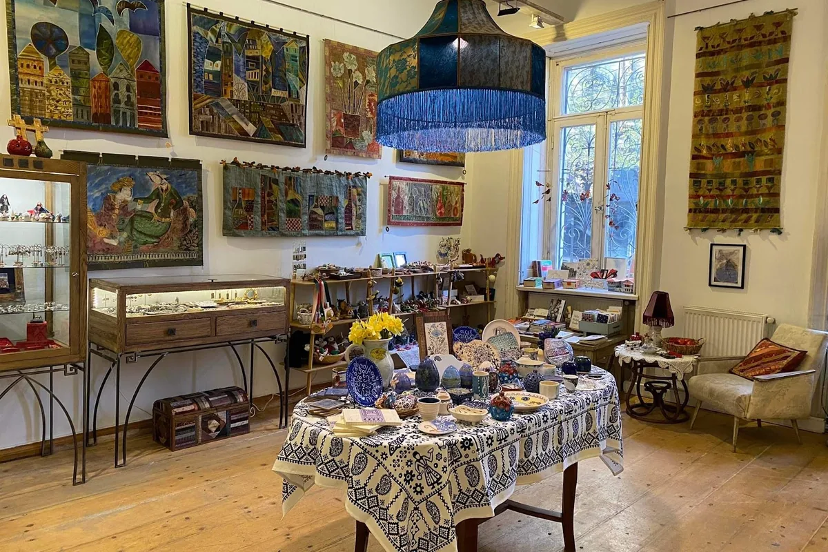 A round table of blue painted ceramics on a white cloth with a dark blue print, glass jewelry cases and fabric wall hangings inside Gallery 27 on Lado Asatiani Street