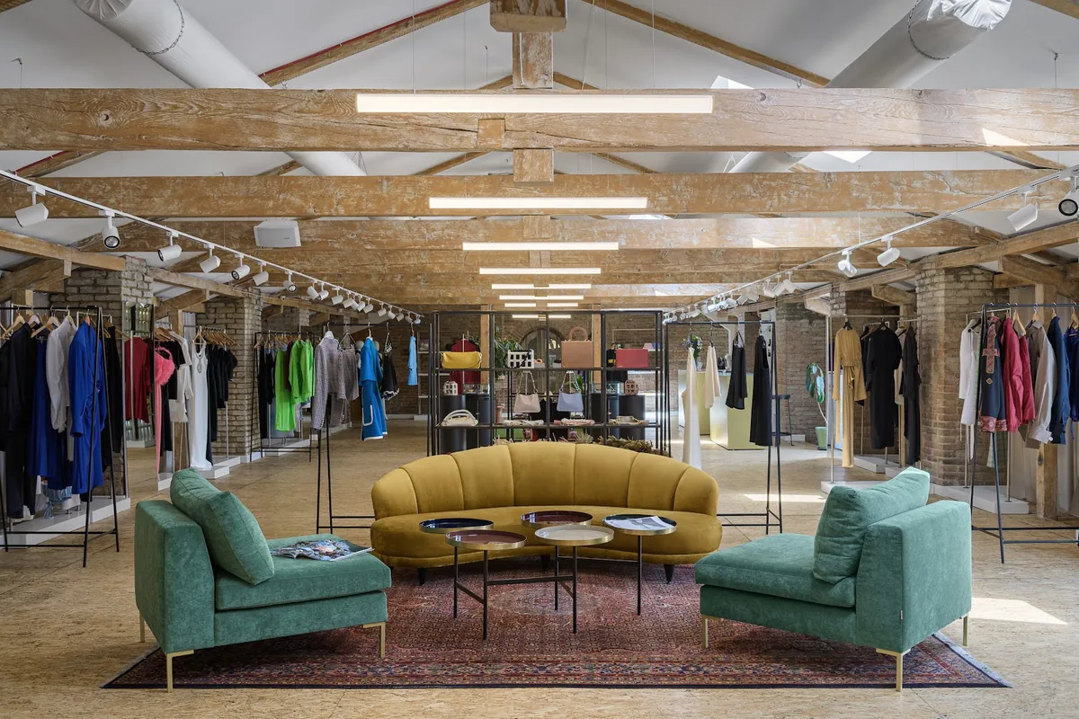 Clothing rails, a curved mustard sofa and green armchairs under old timber roof beams at IERI