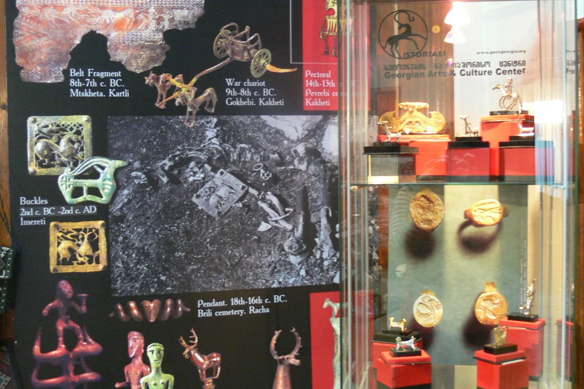 Small gold and silver replica figurines and a gold ornament on red stands in a glass case in front of the Istoriali logo, beside a poster of ancient Georgian metalwork, in the showroom on Niko Nikoladze Street