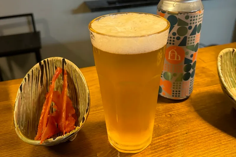 Glass of beer beside a craft beer can and a dried fish snack at Optimist Craft Bar