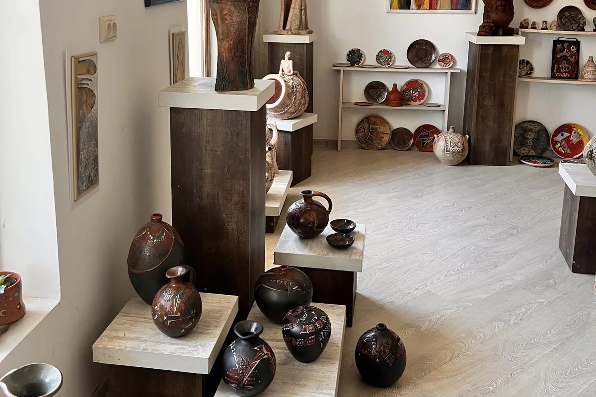 Black-smoked jugs and vases on dark wooden plinths with pale tops in the showroom at ORKOL Studio in Zugdidi, with clay sculptures and painted plates behind