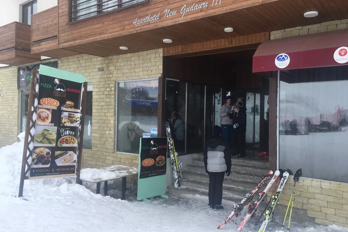 Posticino's pizza and burger signs beside the snowy entrance steps, with skis leaning on the wall, at Posticino in Gudauri