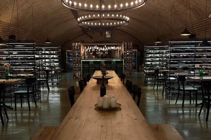 Vaulted brick cellar with a long wooden communal table and chandeliers at Sadzvele Wine Hub in Wine Factory N1