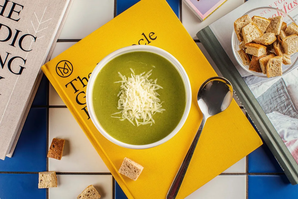Green soup with grated cheese and croutons on a stack of books at They Said Books