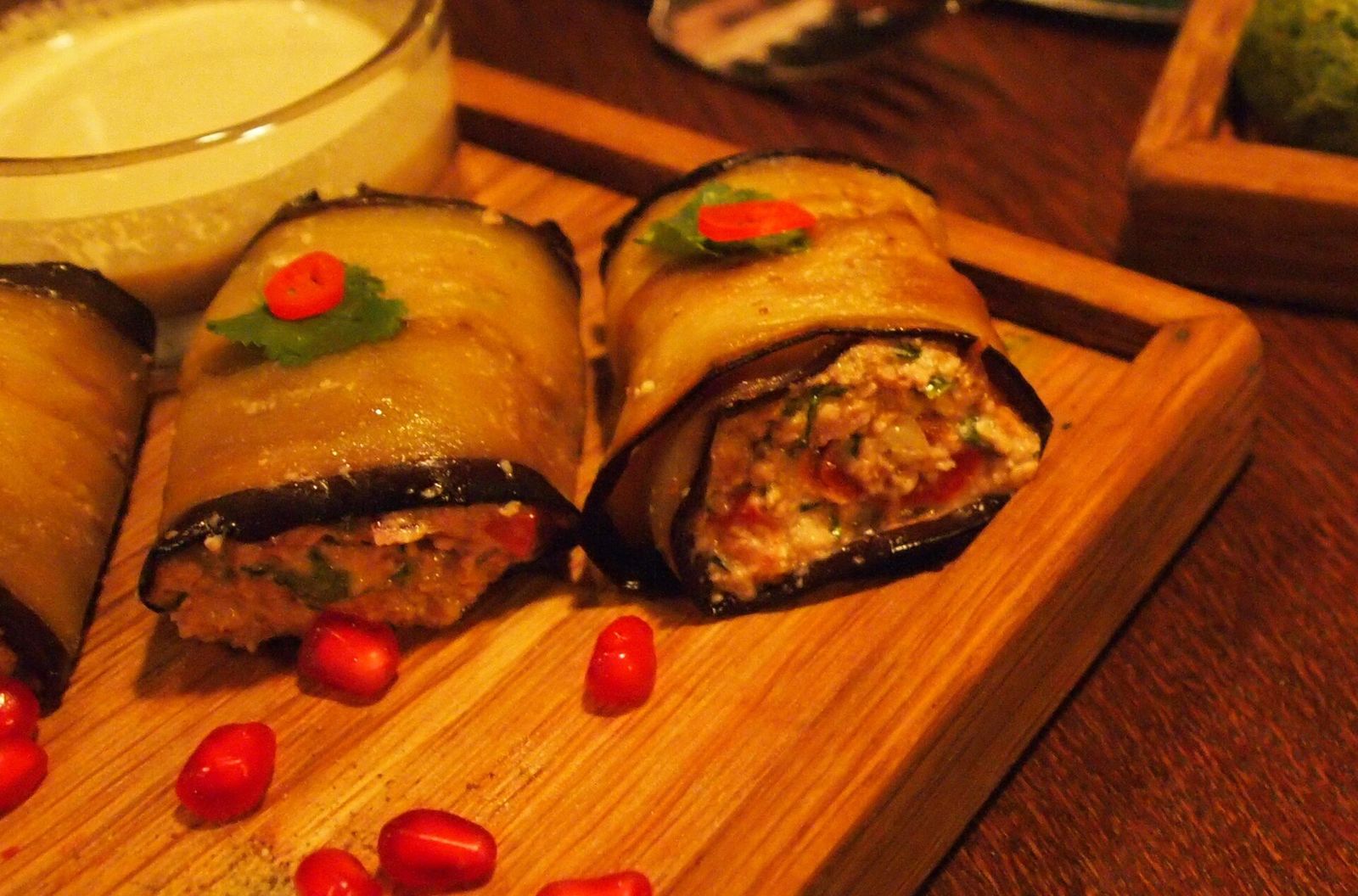 Badrijani nigvzit, fried eggplant slices rolled around walnut paste, each topped with a chilli slice and a herb leaf, with pomegranate seeds scattered on the wooden board