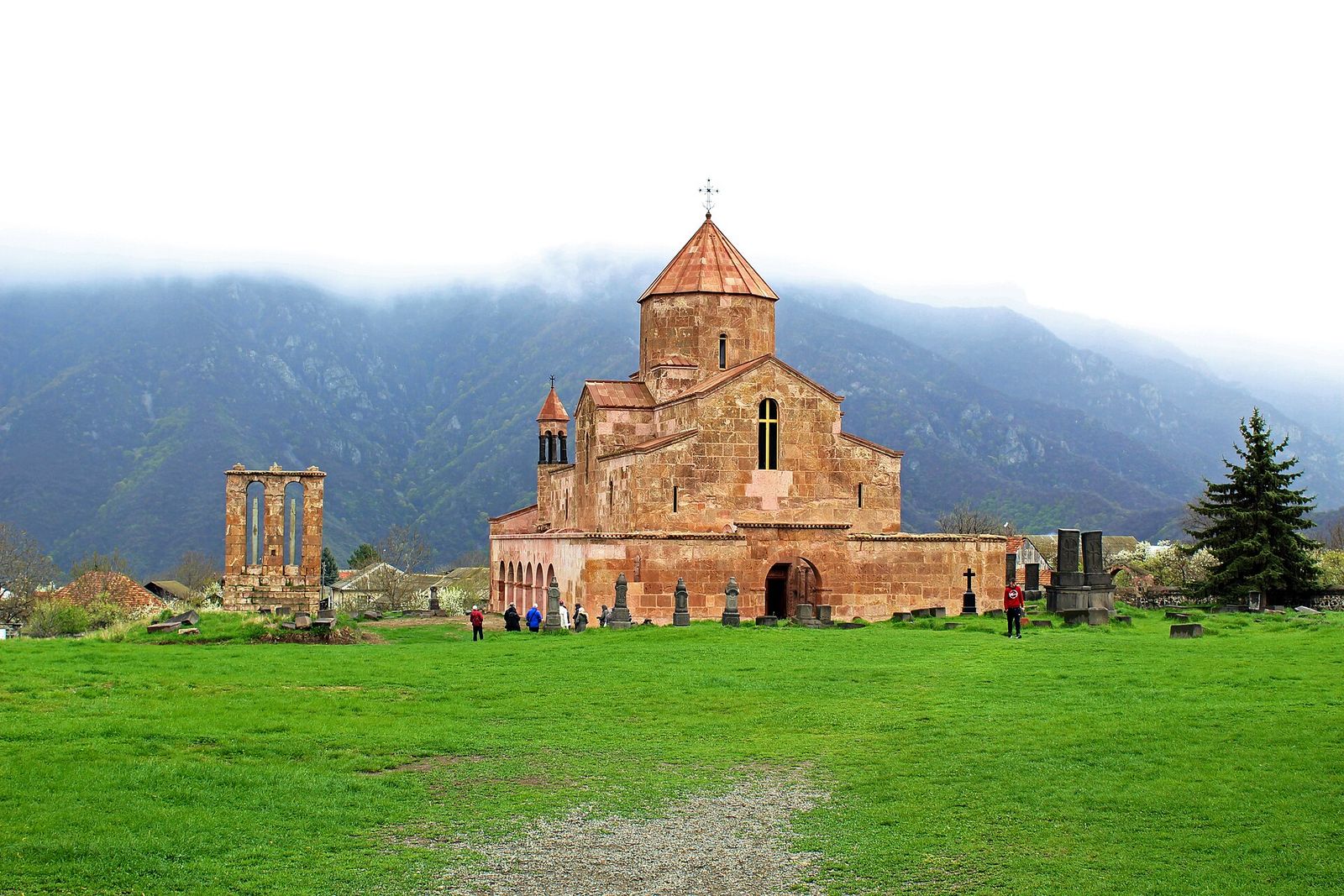 Odzun Church, a domed stone basilica on a green lawn, with its arched funerary monument to the left