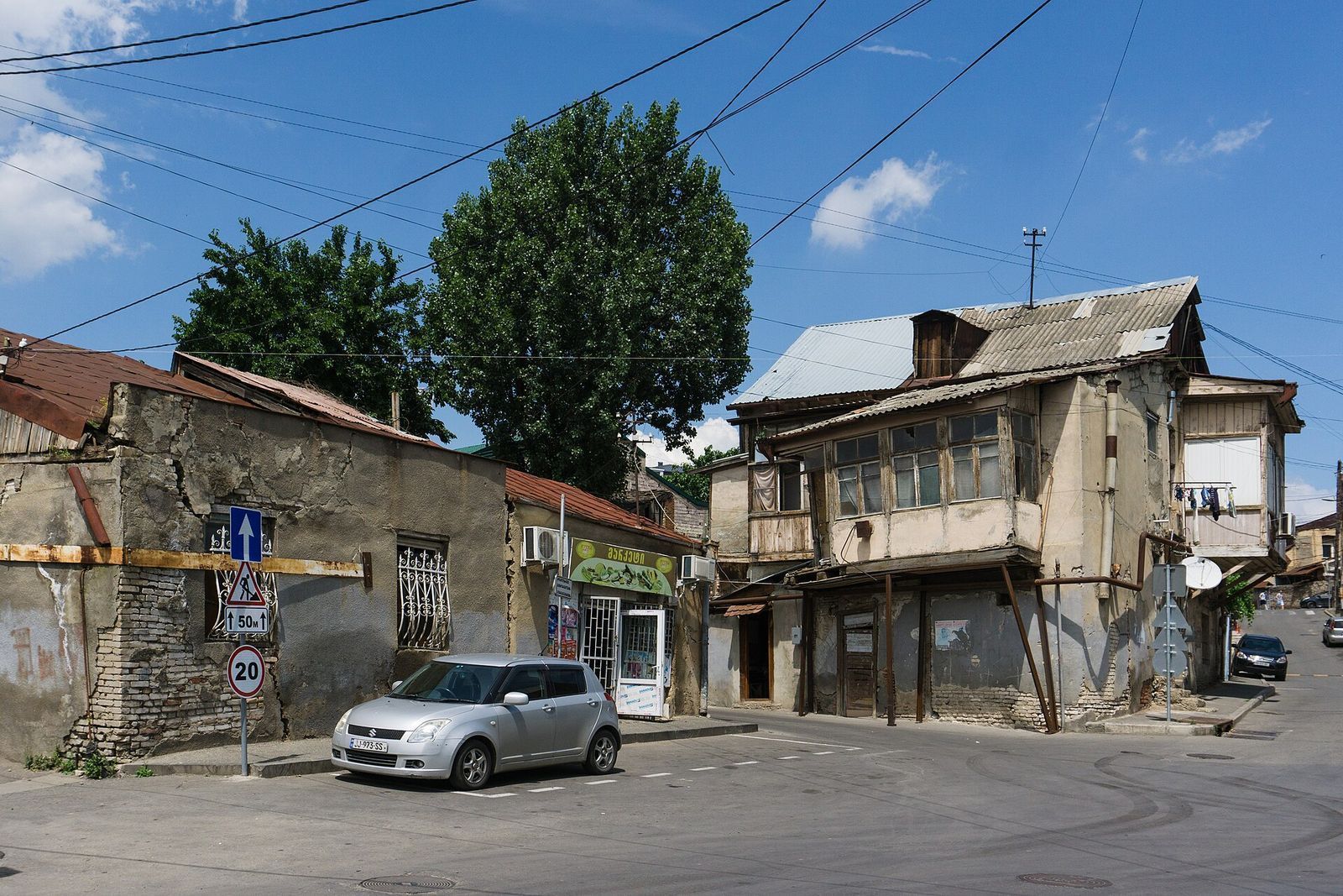 An Avlabari street corner with an old house and glassed-in wooden balcony, a corner shop and a parked car
