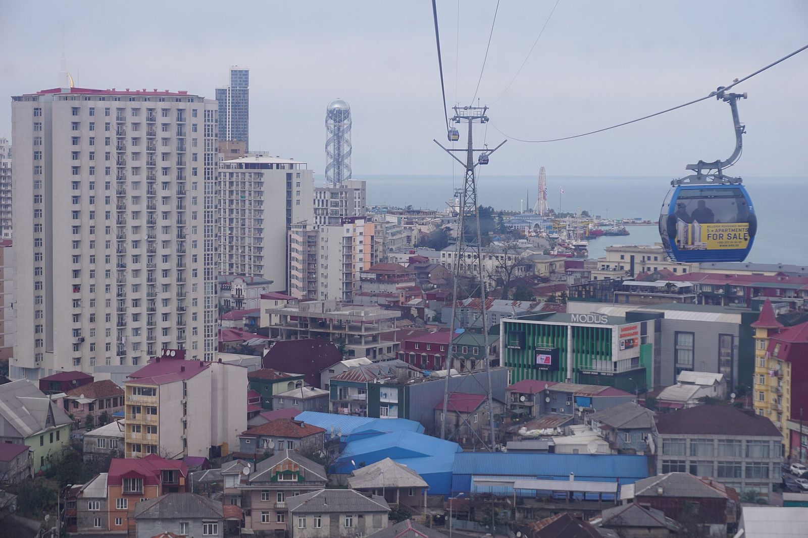 A blue Argo cable car gondola above Batumi, with the Alphabet Tower and the Black Sea behind
