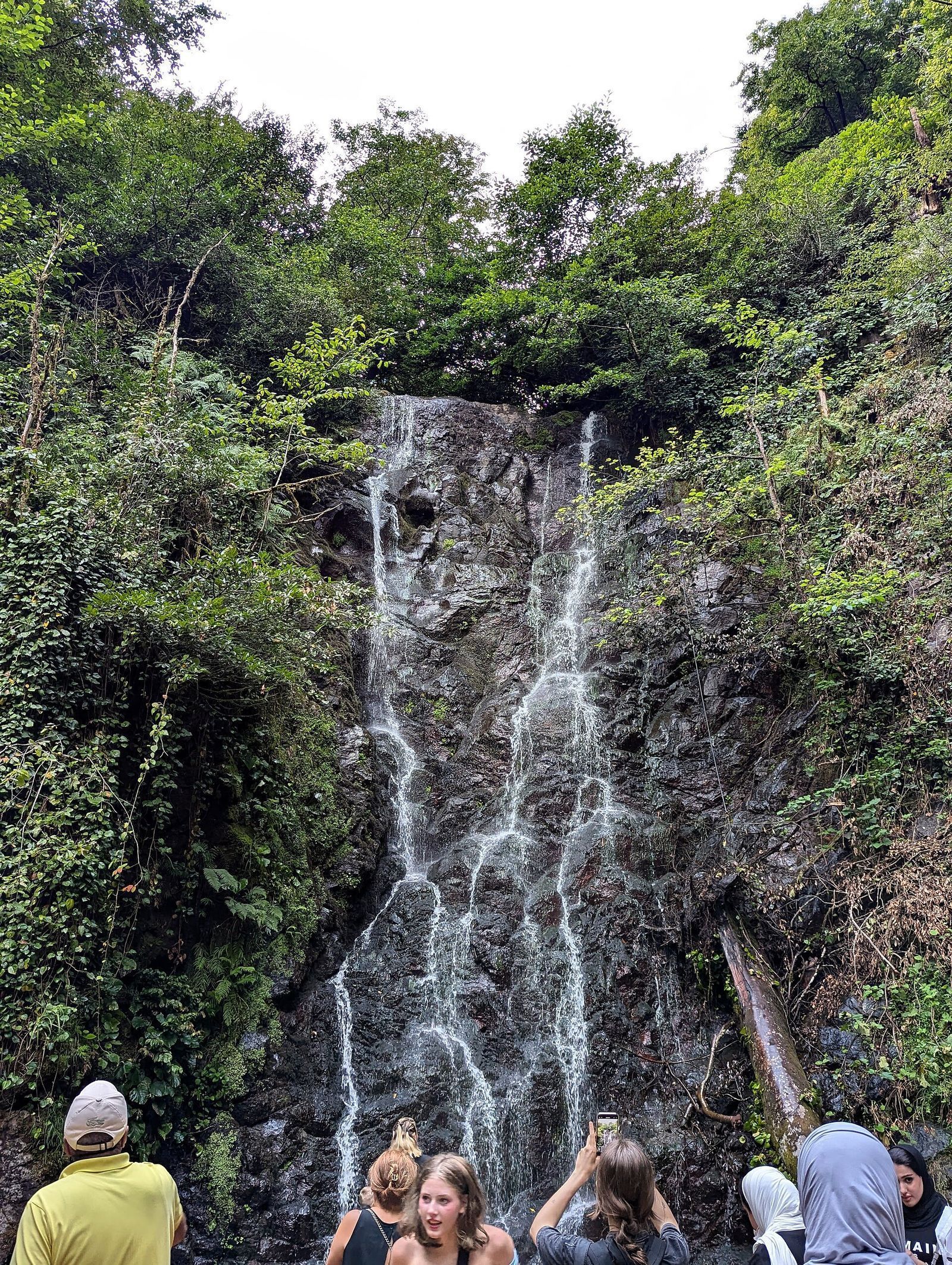 Mirveti waterfall running in thin streams down a dark rock face in green forest, with visitors in front