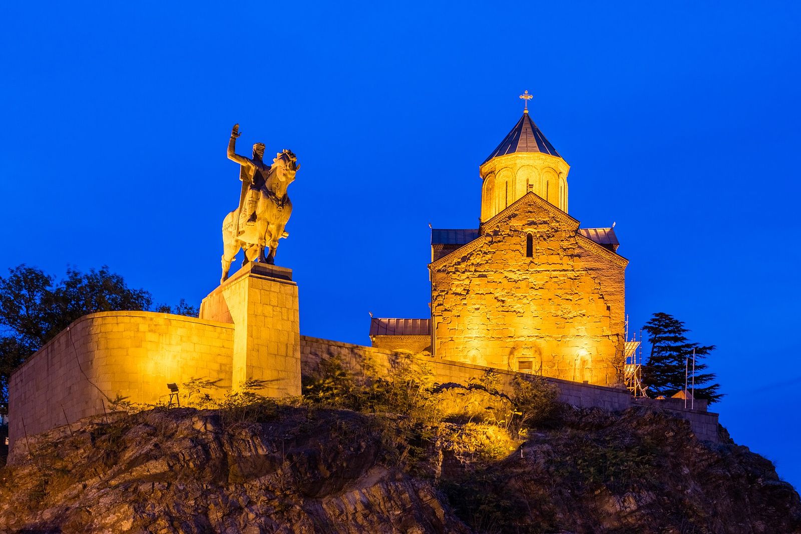 Metekhi Church floodlit at night on its rock above the river, with the statue of King Vakhtang Gorgasali on horseback beside it
