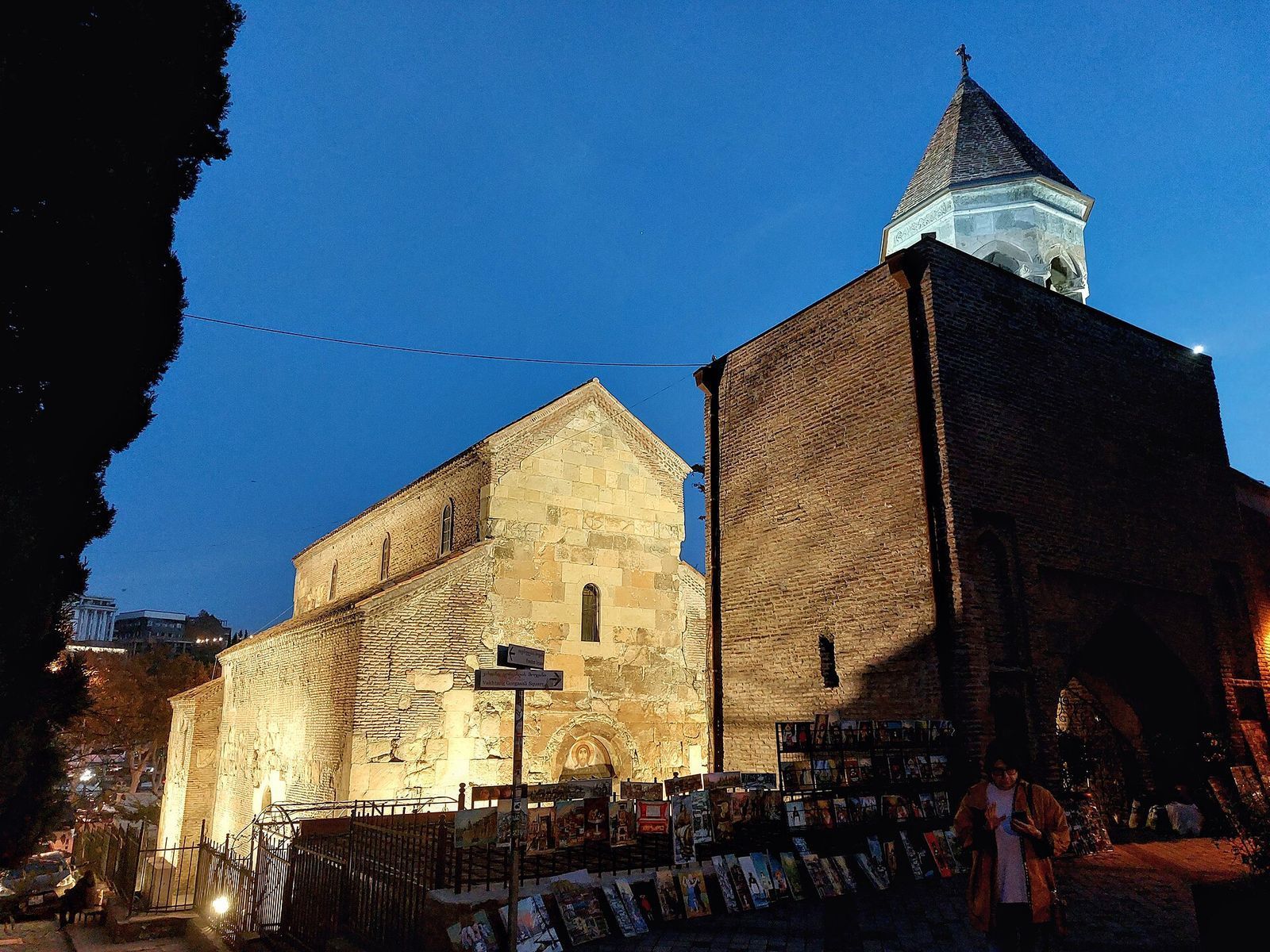 The stone walls of Anchiskhati Basilica and its brick bell tower at dusk, with paintings for sale along the railings