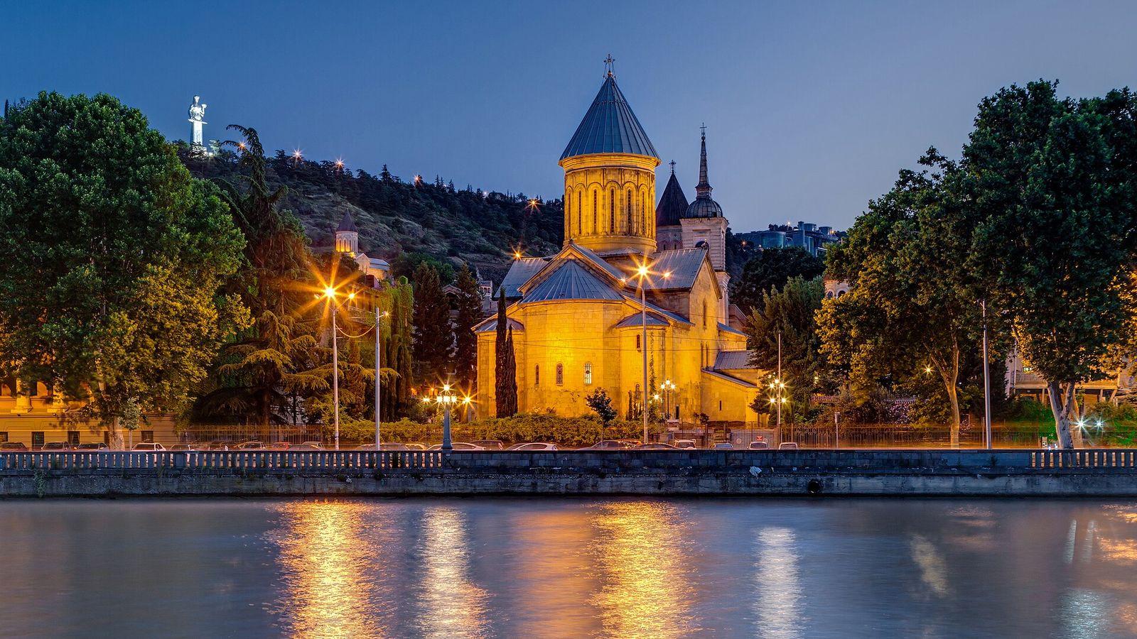 Sioni Cathedral lit up at dusk on the bank of the Mtkvari, with the Mother of Georgia statue on the ridge behind