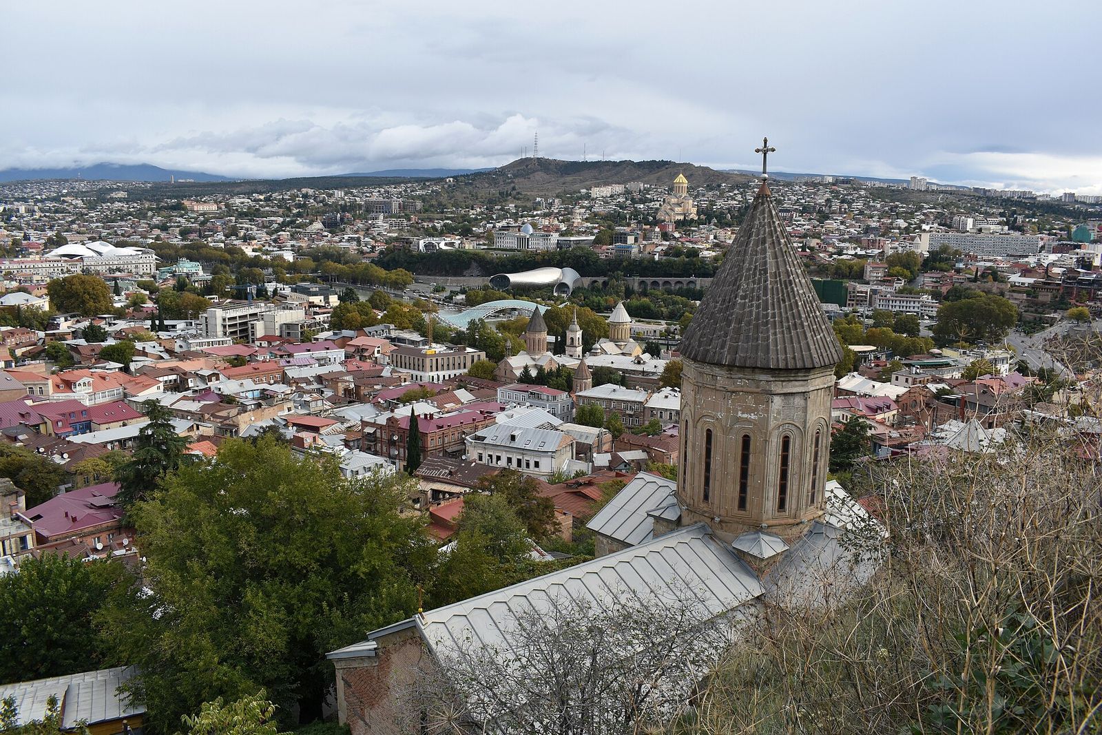 The stone drum and conical dome of Upper Betlemi Church above the Old Town rooftops, with Sameba's gold dome on the hill across the river