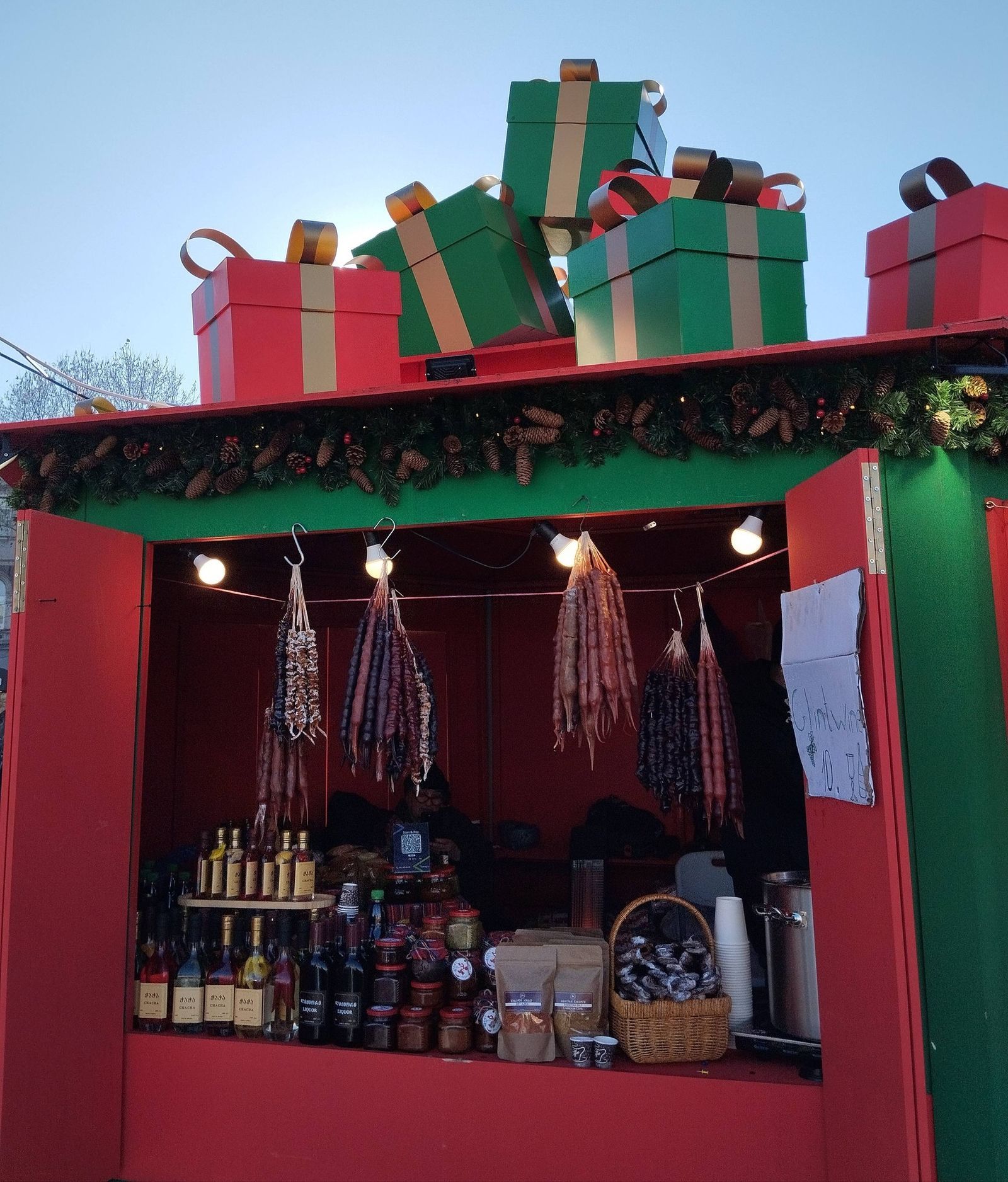 Red and green holiday market stall in Tbilisi hung with churchkhela, with bottles of chacha and a mulled wine sign