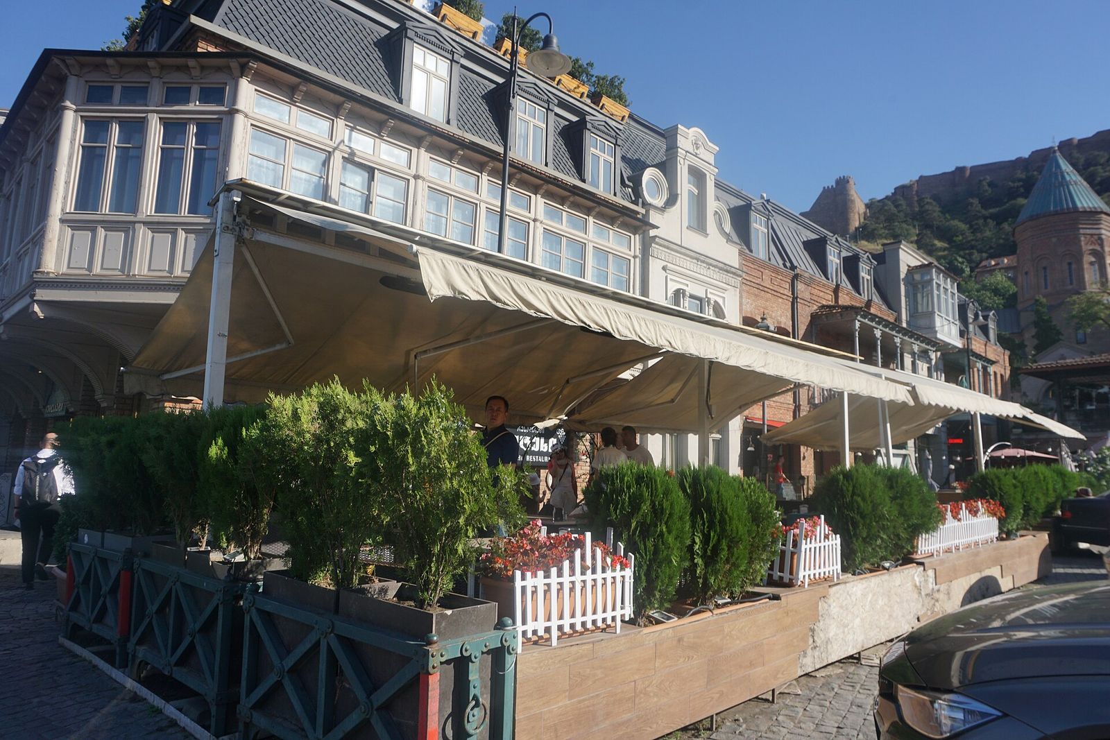 Restaurant terraces under awnings in Old Tbilisi, with the walls of Narikala Fortress on the ridge above