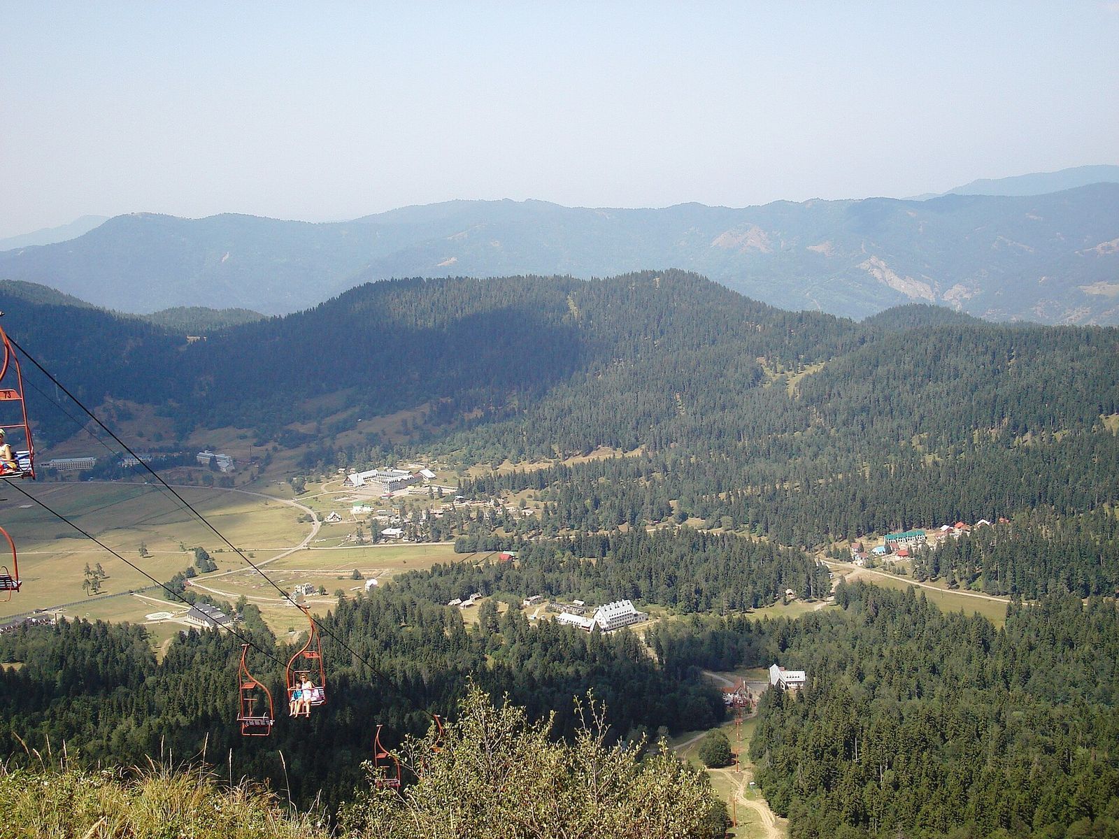 Bakuriani seen from Mount Kokhta: the resort spread across a green valley between forested hills, with chairlift seats in the foreground