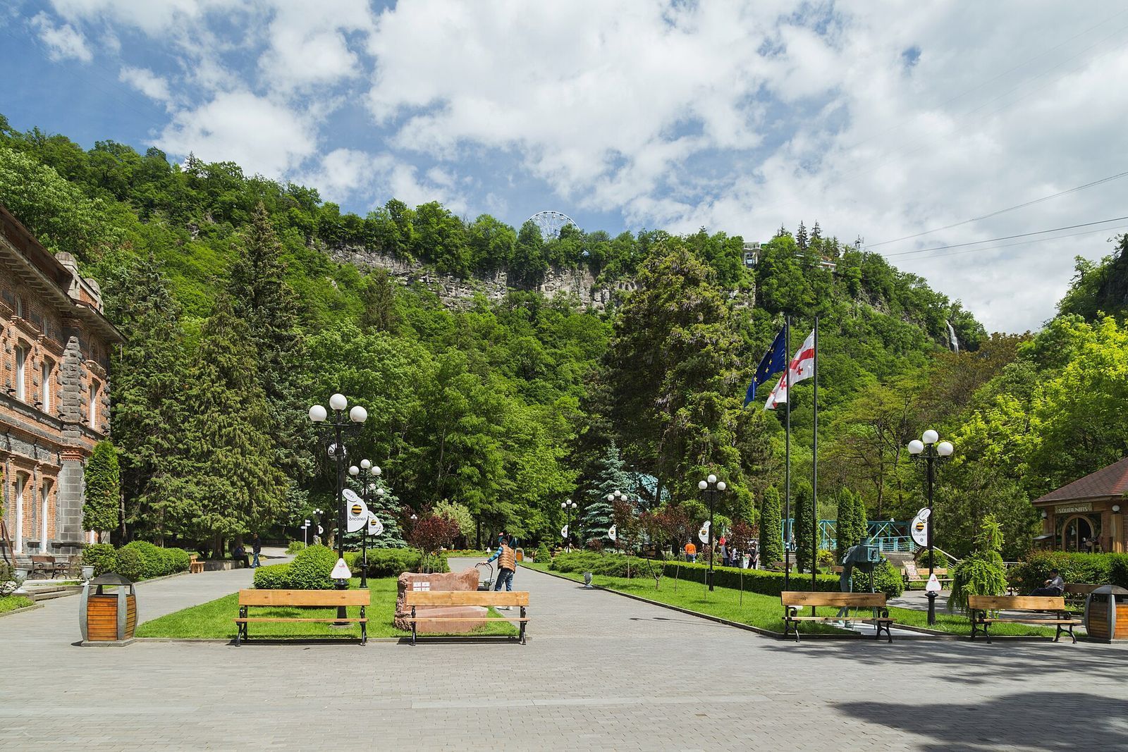 The entrance square of Borjomi Central Park, with benches, lamps and flags in front of the forested cliffs of the gorge