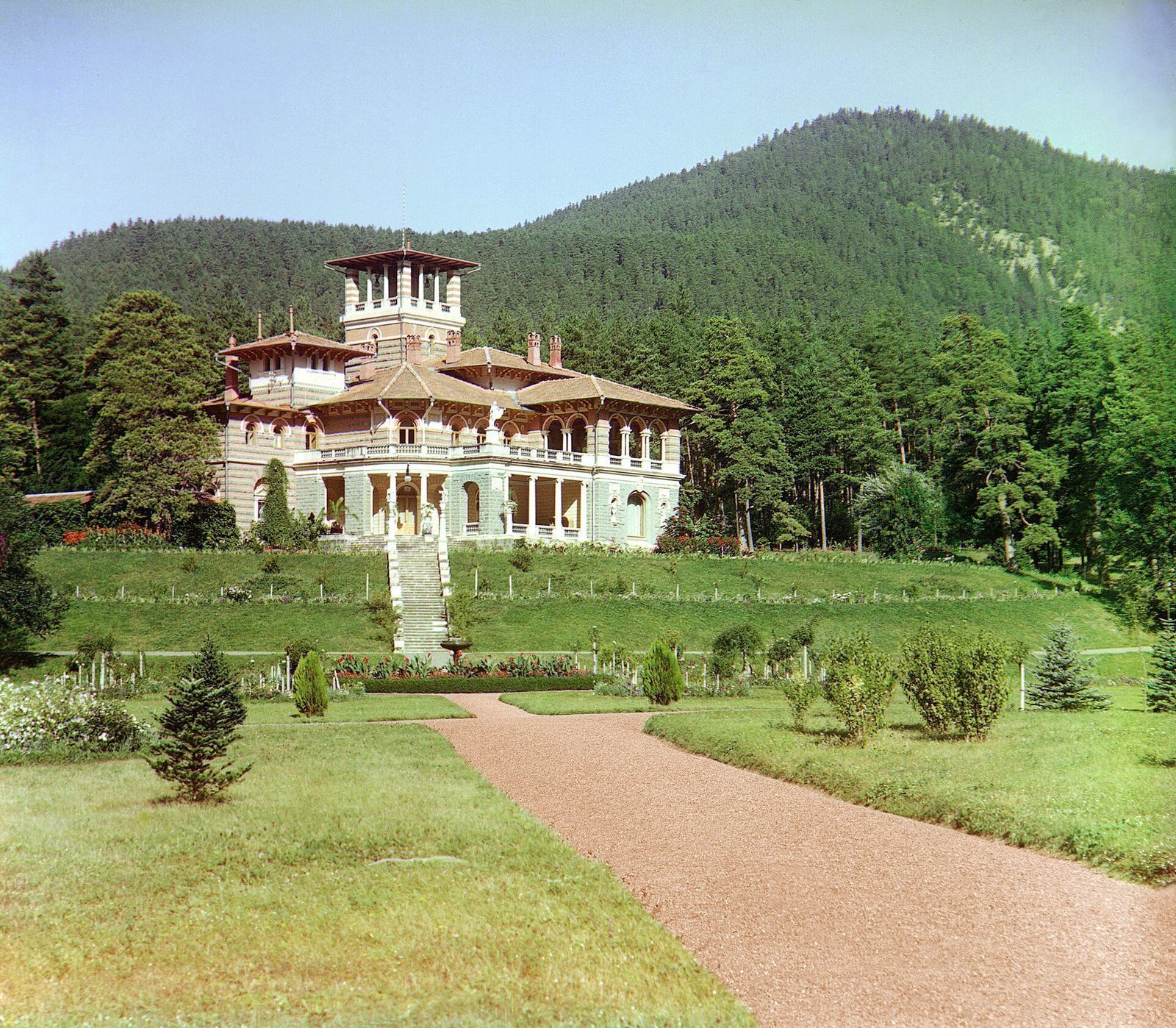 Likani Palace in Borjomi in an early color photograph, the towered villa above a lawn and gravel path with pine forest behind