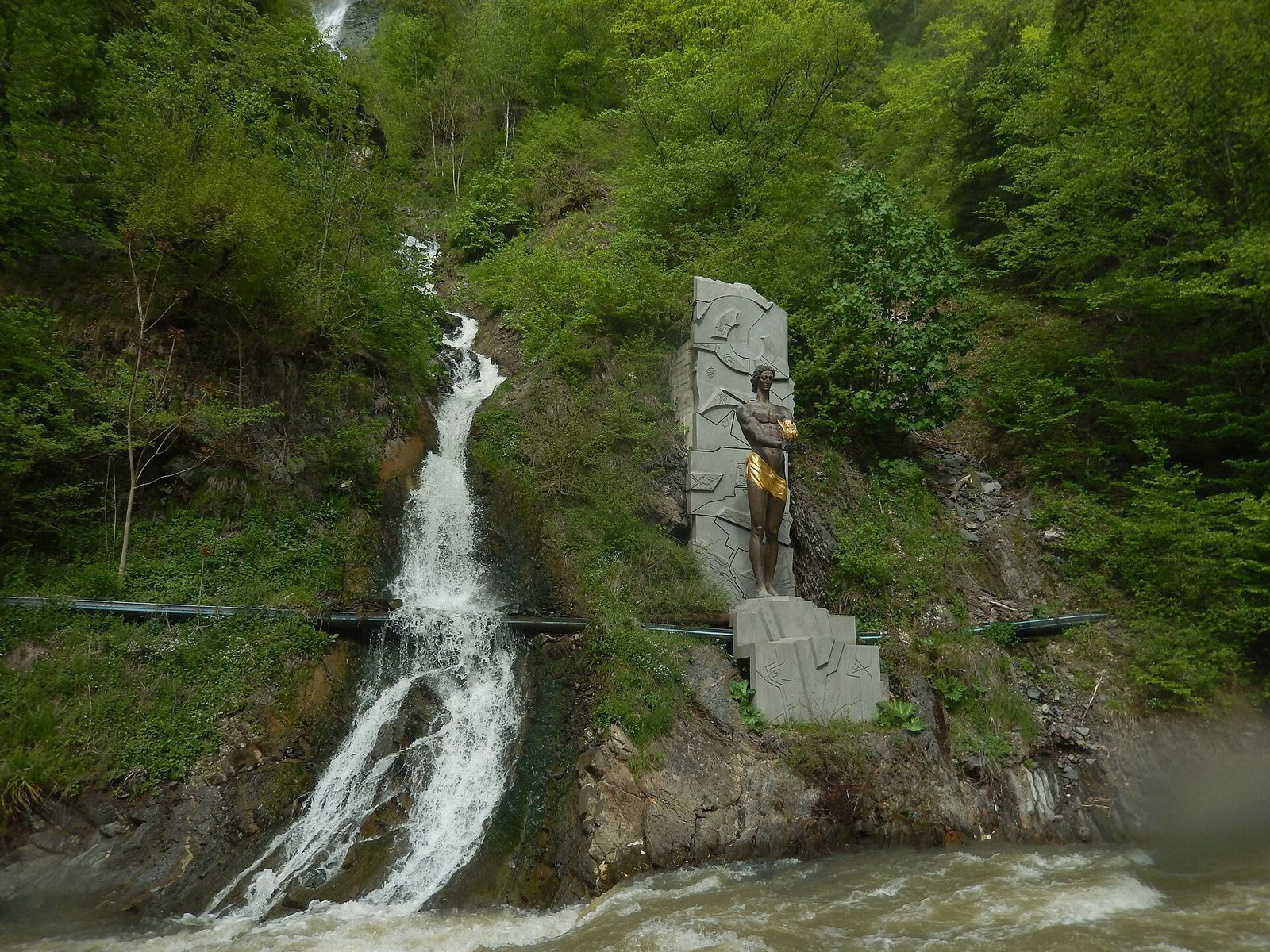 The bronze Prometheus statue in Borjomi Central Park, holding a gilded flame in front of a carved stone slab, beside a waterfall that runs down the wooded slope into the Borjomula