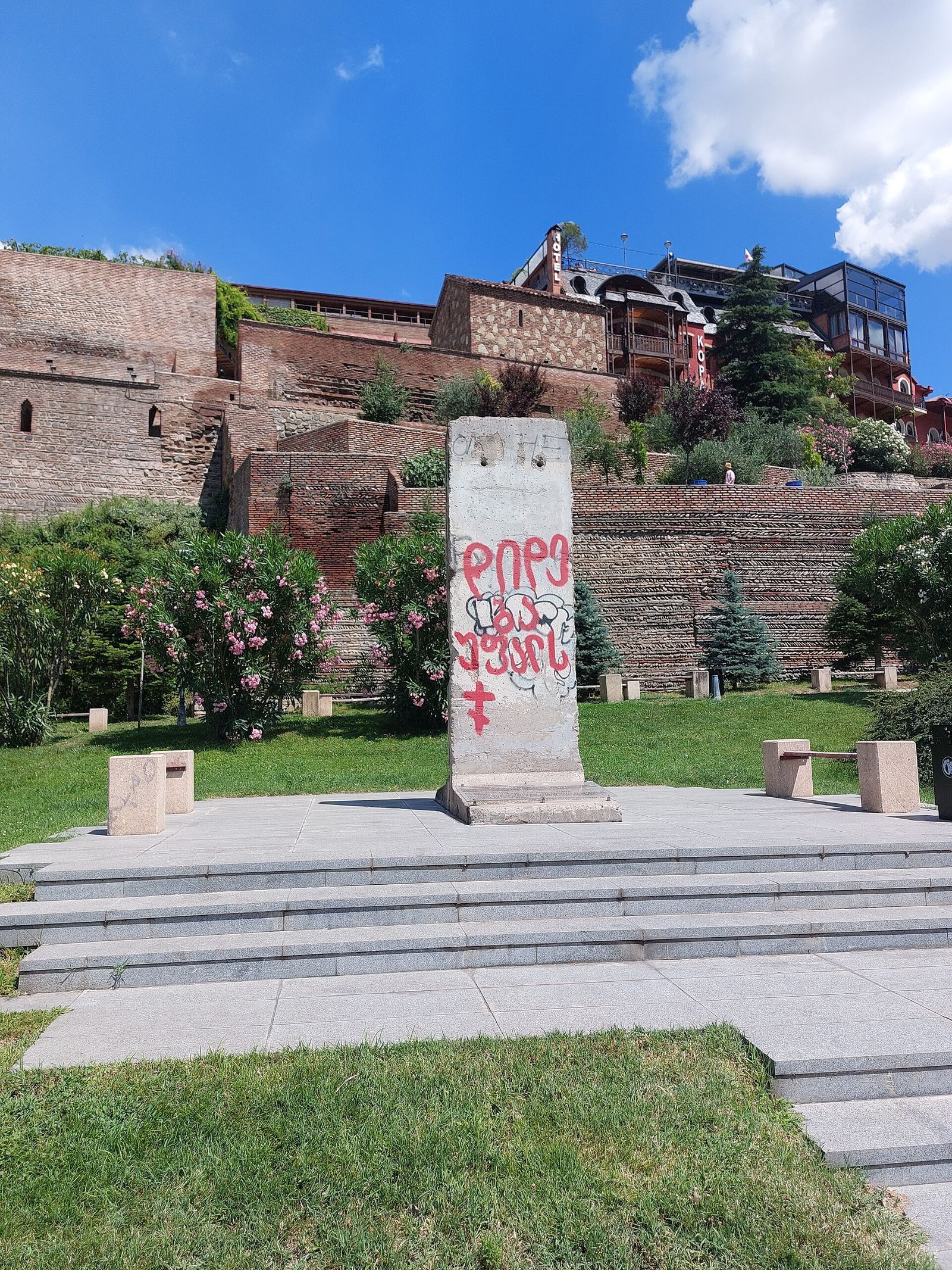 The Berlin Wall segment on its plinth in Europe Square, Tbilisi, with red Georgian graffiti and the brick terraces of Metekhi behind