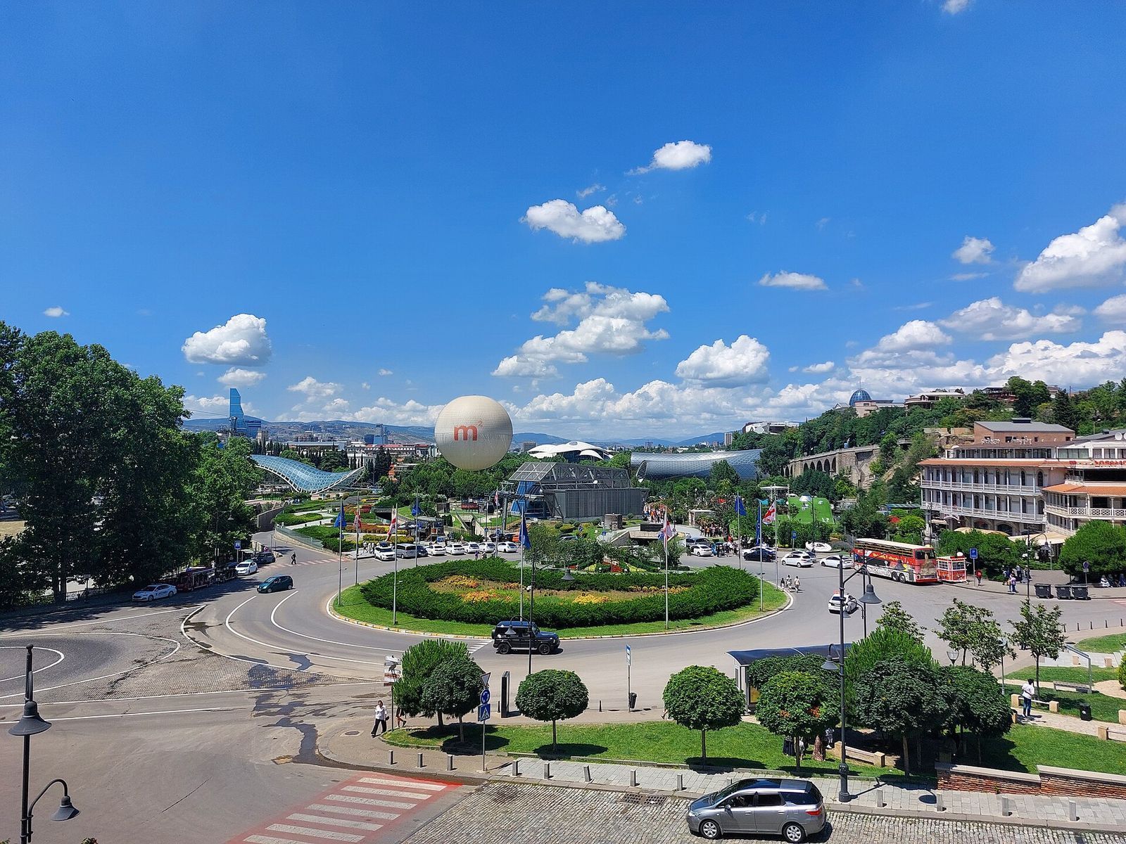 Europe Square in Tbilisi with flower beds and flags, the Rike Park tubes, the Bridge of Peace canopy and a tethered balloon behind