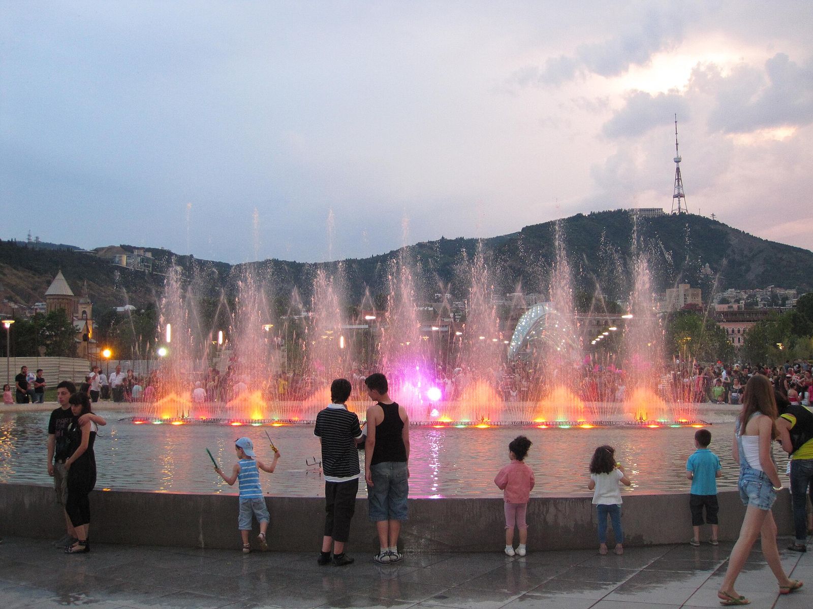 Children watching the lit-up musical fountain in Rike Park at dusk, with the Bridge of Peace and Mtatsminda behind
