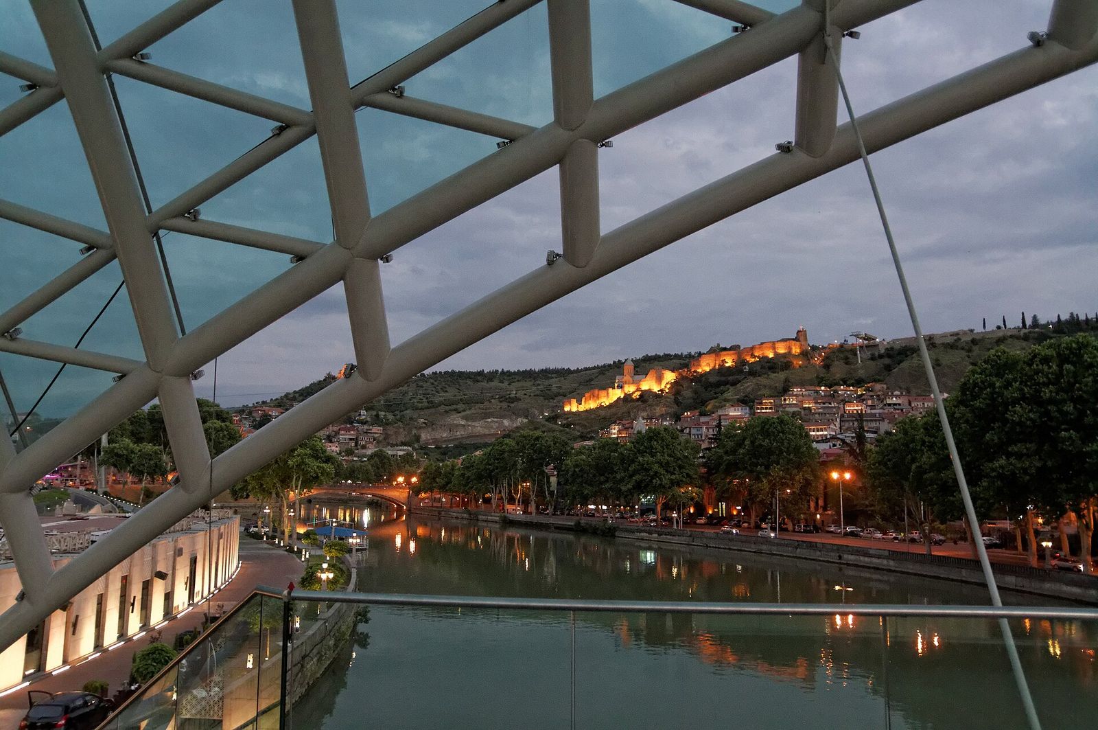 The view through the steel ribs of the Bridge of Peace at dusk toward the floodlit walls of Narikala Fortress above the Kura river