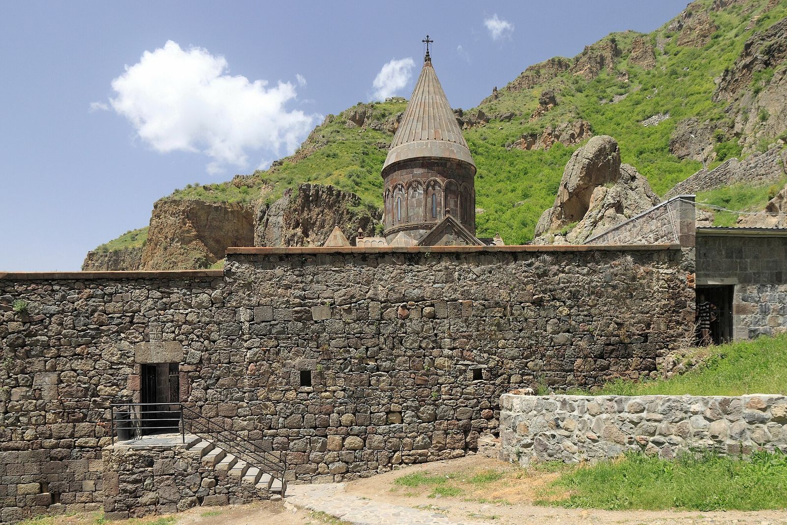 The stone dome of Geghard Monastery behind its outer wall, with green cliffs above, in Armenia