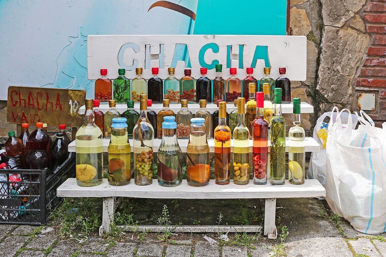 Bottles of homemade fruit chacha with whole fruit inside, lined up on a white bench under a CHACHA sign at a stall in Mtskheta