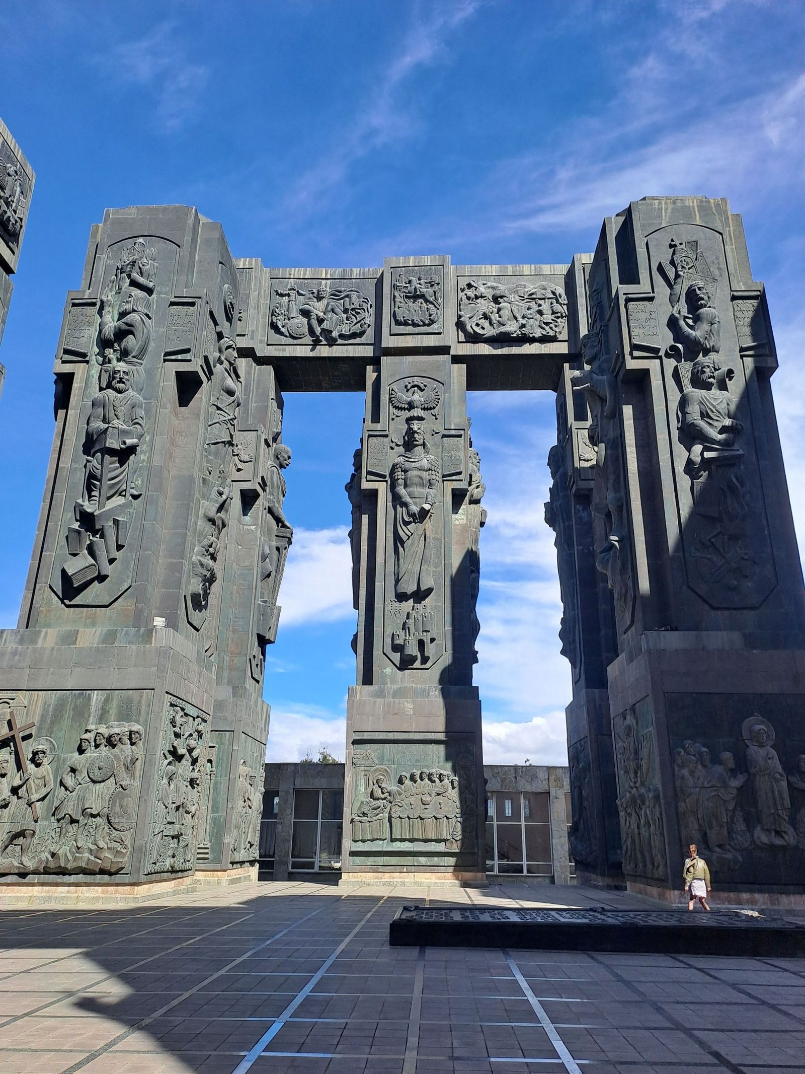 The tall carved pillars of the Chronicles of Georgia around the monument's paved square, with a visitor at their foot