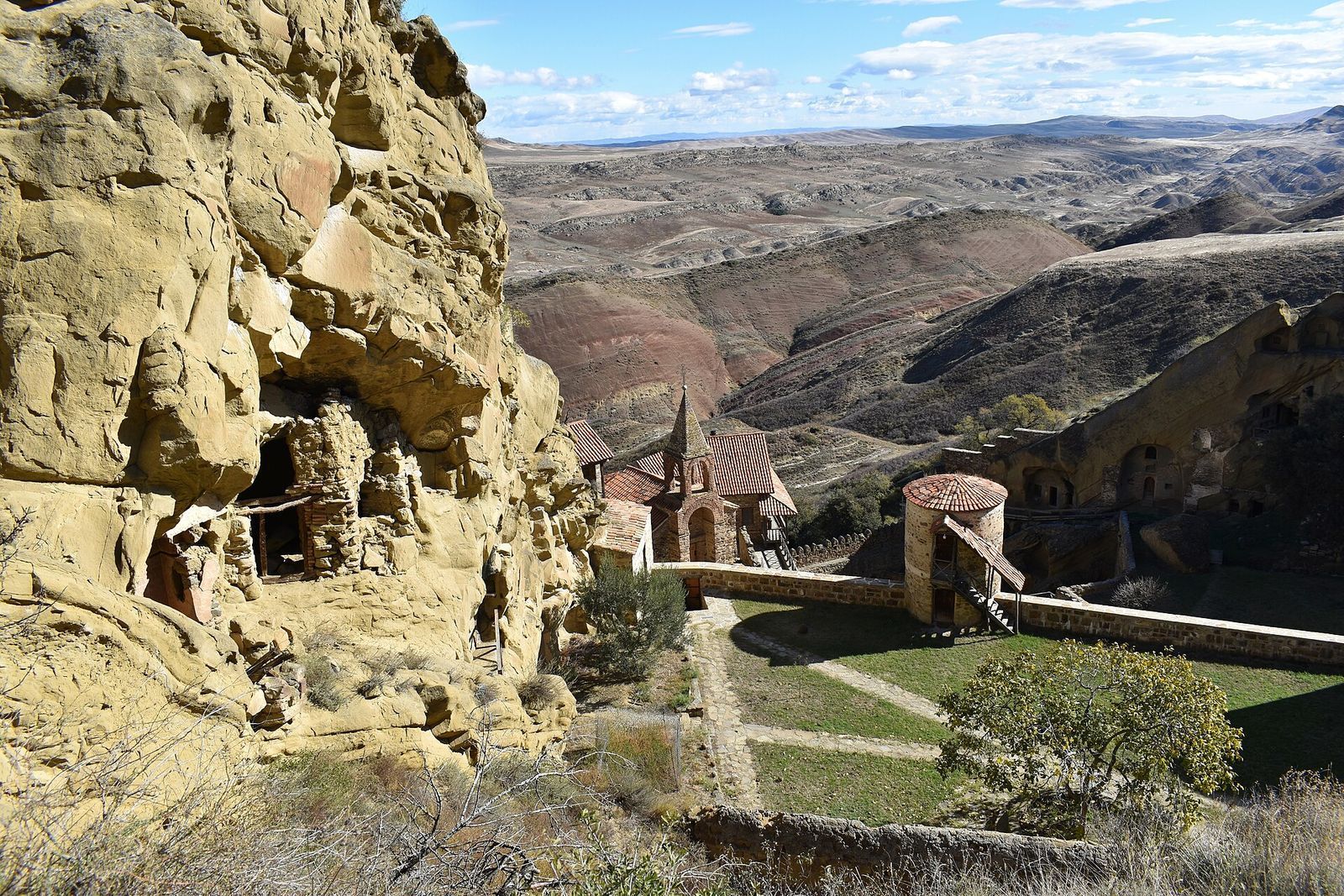 Lavra Monastery at David Gareja from above: cave cells in the sandstone cliff, a church with a red tiled roof, a round tower and the striped desert hills beyond
