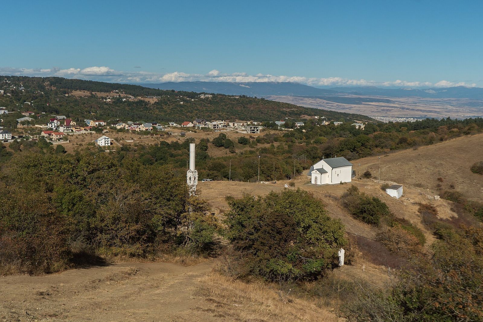 The houses of Kojori on the hilltop, with a small white church on the grassy slope below, seen from Kojori Fortress