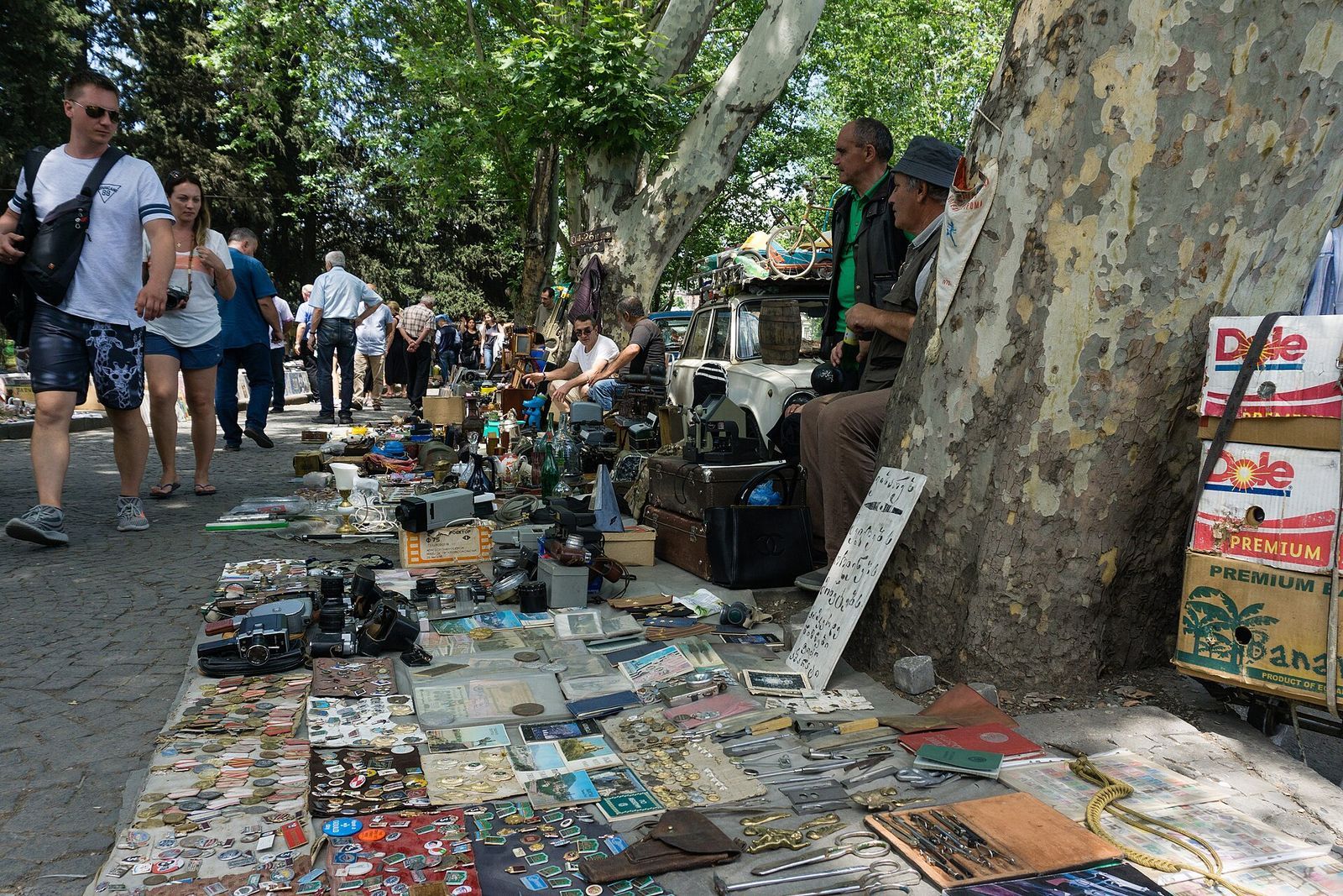 Soviet badges, coins, postcards and old cameras laid out on the pavement under the plane trees at the Dry Bridge flea market, with browsers walking past