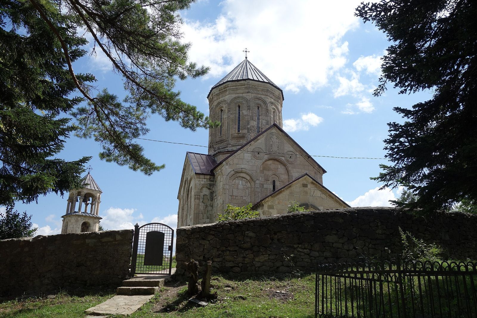 Nikortsminda Cathedral in Racha, its carved stone drum and separate bell tower behind a stone wall among fir trees