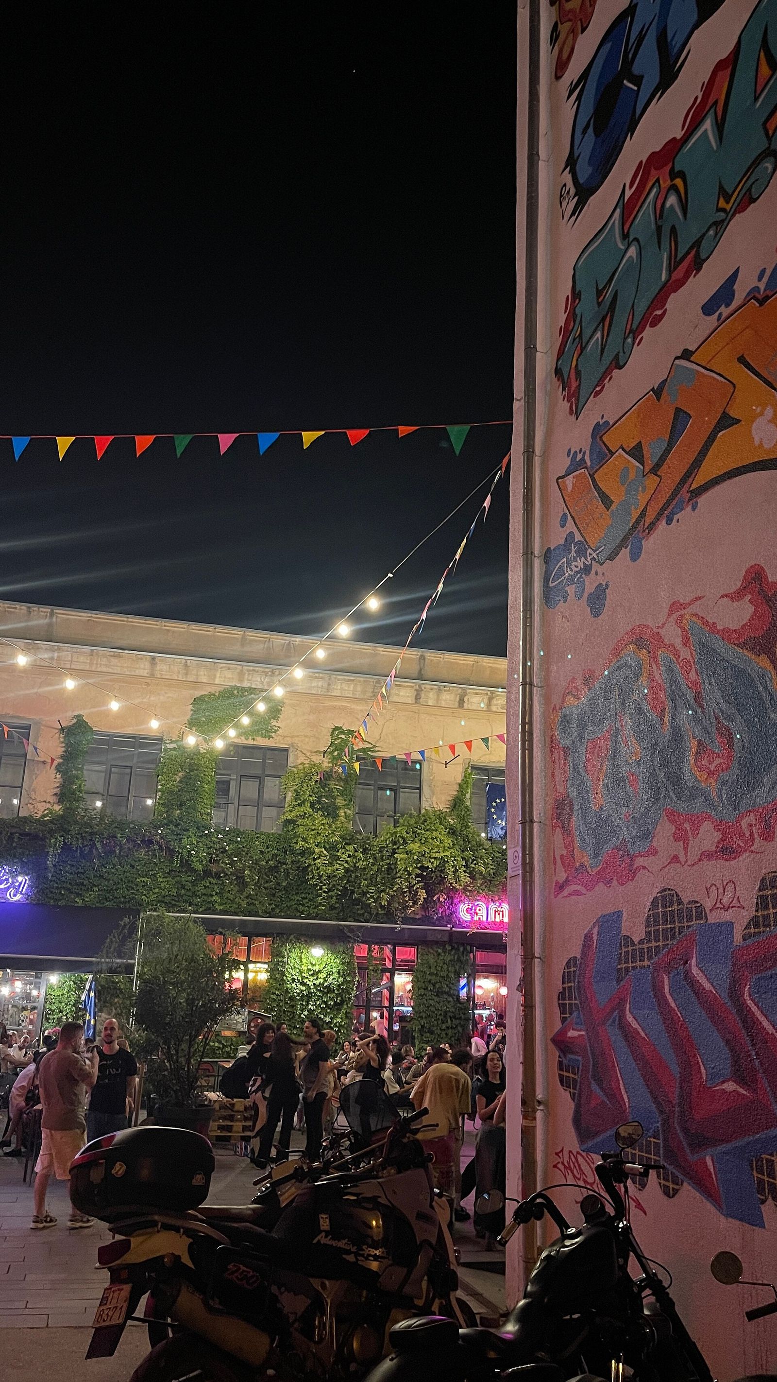 Graffiti covering a wall of the Fabrika courtyard at night, with string lights and a crowd at the bar tables behind