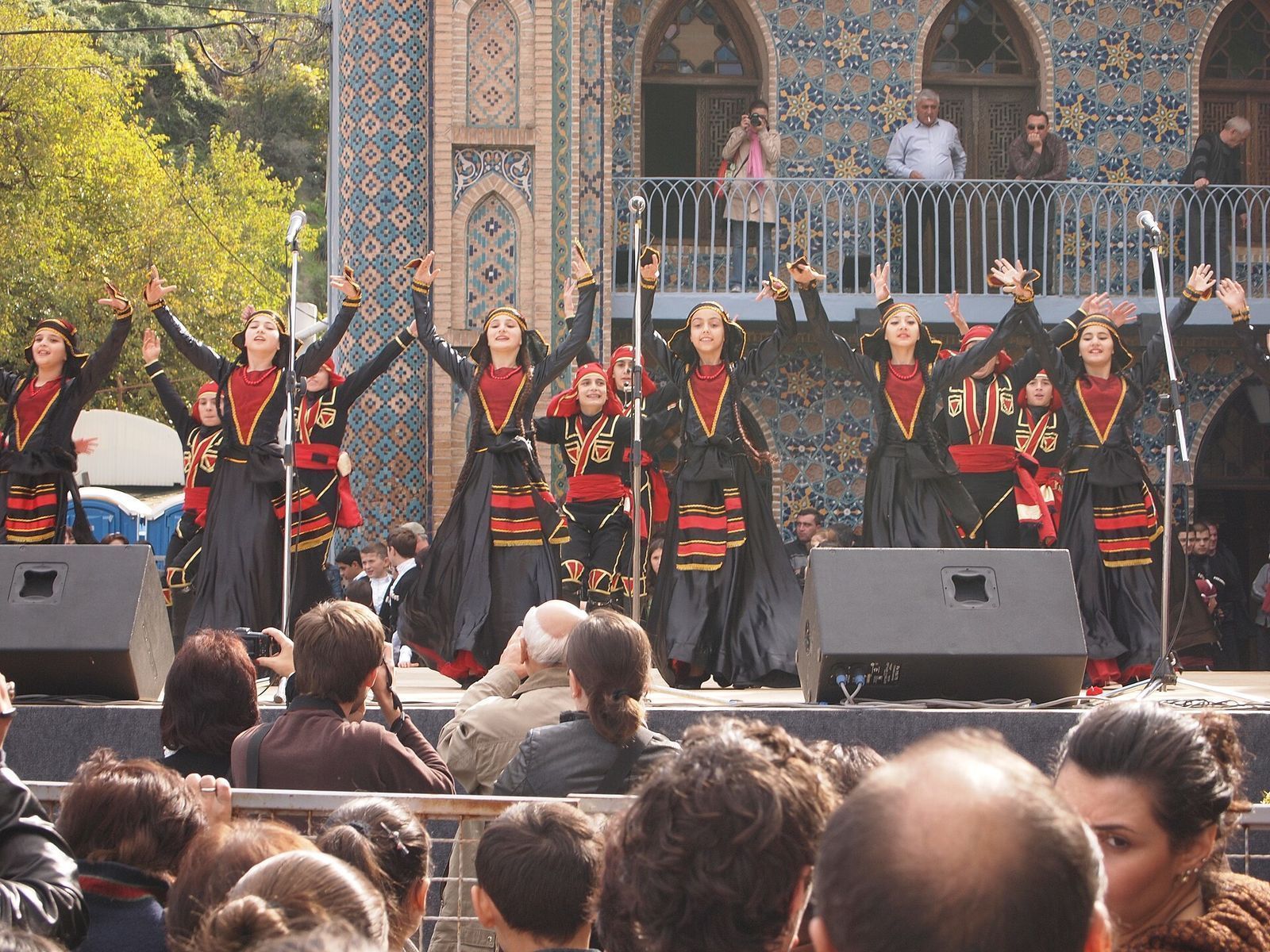 Young dancers in black and red folk costume on a Tbilisoba stage in front of the blue-tiled Orbeliani bathhouse in Abanotubani