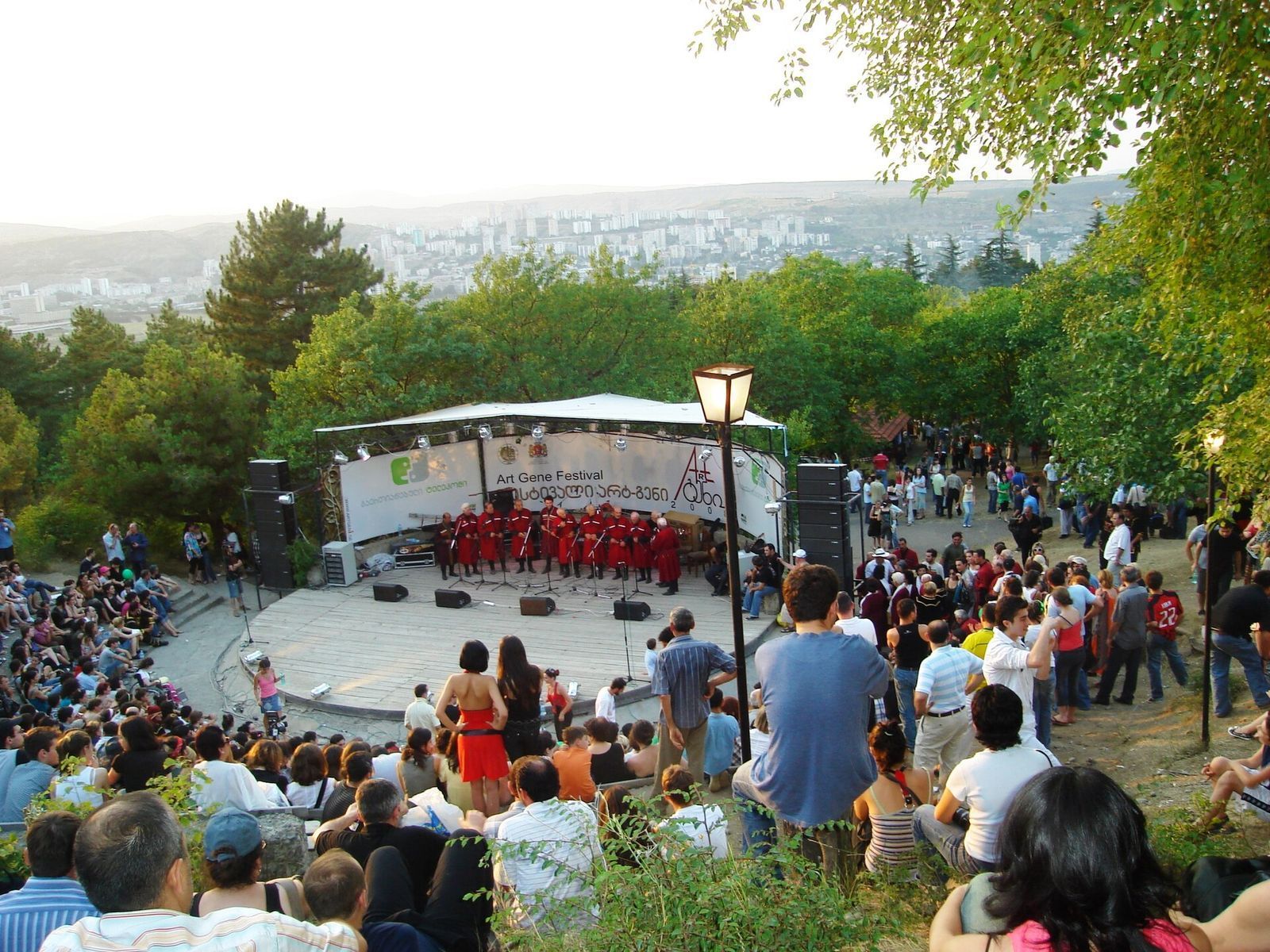 A folk ensemble from Guria in red costume on the Art-Gene stage at the Open Air Museum of Ethnography, with the audience on the slope and Tbilisi behind