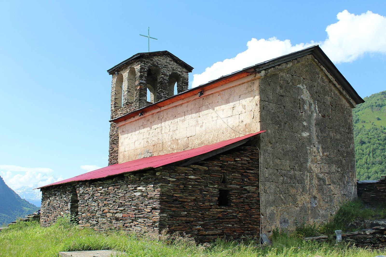 The stone Lagurka church of St Kvirike and St Ivlita in Kala, Svaneti, with its bell tower and red tin roof against the mountains