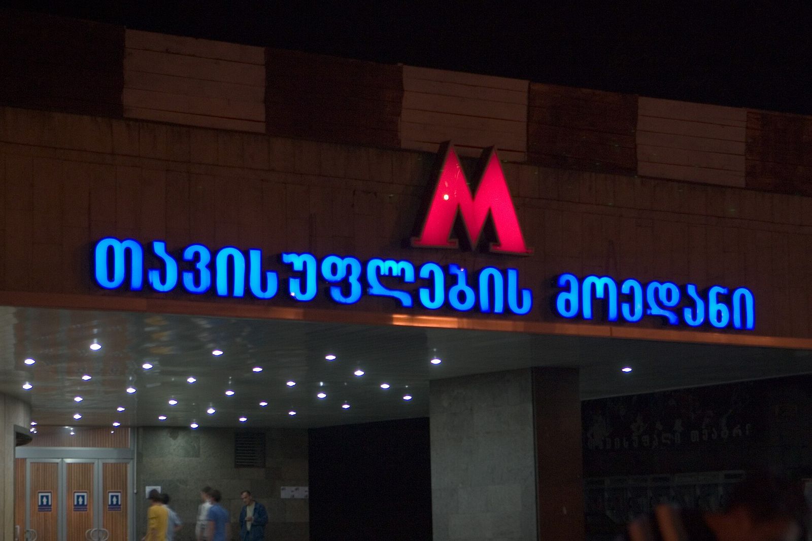 The entrance of Liberty Square metro station in Tbilisi at night, its red M and blue Georgian lettering lit up above the doors