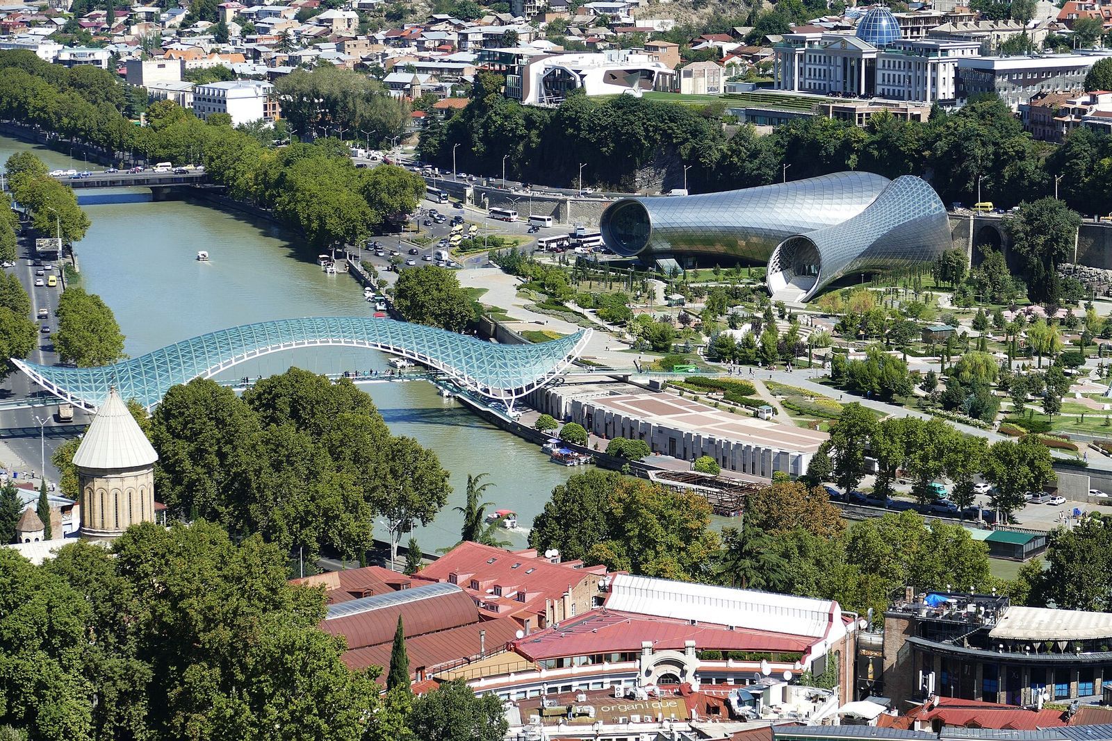 The glass Bridge of Peace landing in Rike Park on the Avlabari bank of the Kura, seen from above with the park's lawns and the Music Theatre tubes