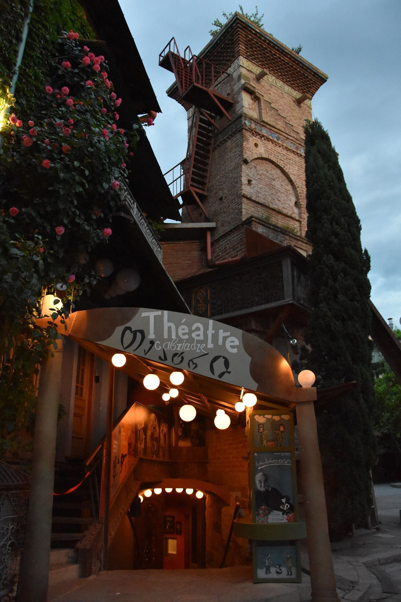 The lit entrance of the Gabriadze Marionette Theatre under its Théâtre Gabriadze sign, with the clock tower rising behind