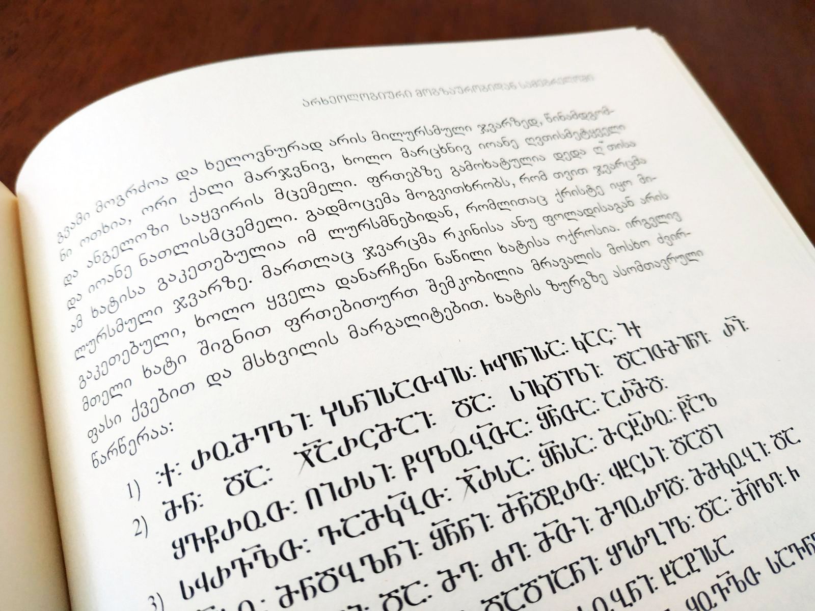 An open Georgian book with a paragraph printed in Mkhedruli and numbered lines in Asomtavruli below it