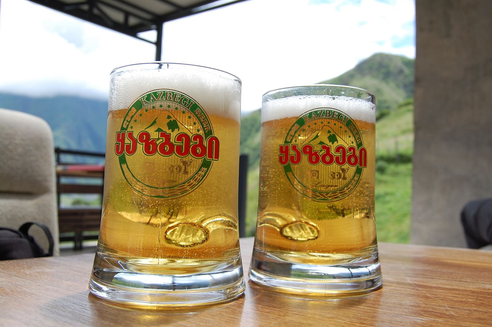 Two branded mugs of draft Kazbegi lager on a café table with green mountains behind in Stepantsminda