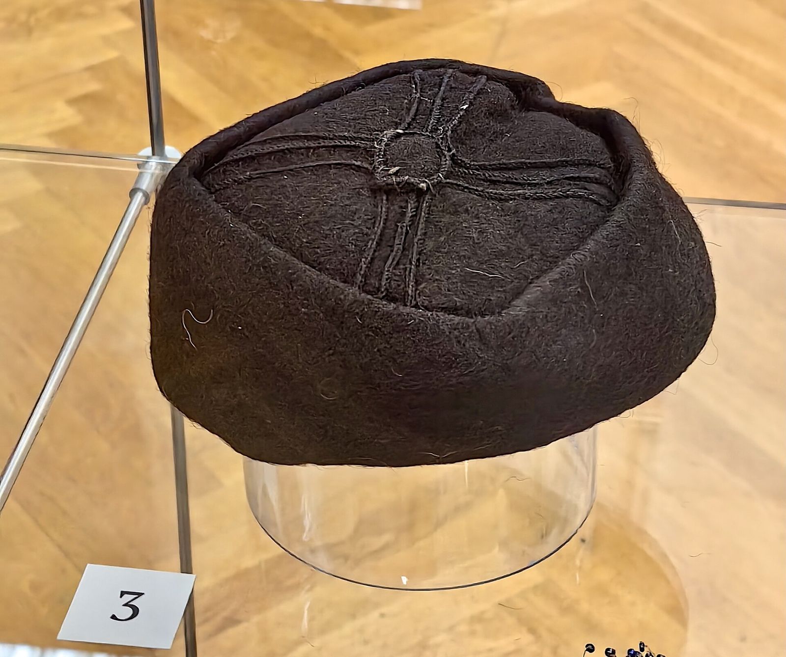A black Georgian felt cap from the early 20th century, a nabdis kudi with stitched lines crossing the crown, on a museum stand